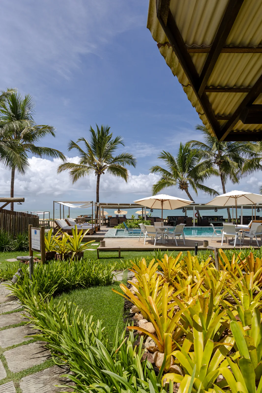 Jardim, piscina e mar vistos do restaurante, com coqueiros e guarda-sóis